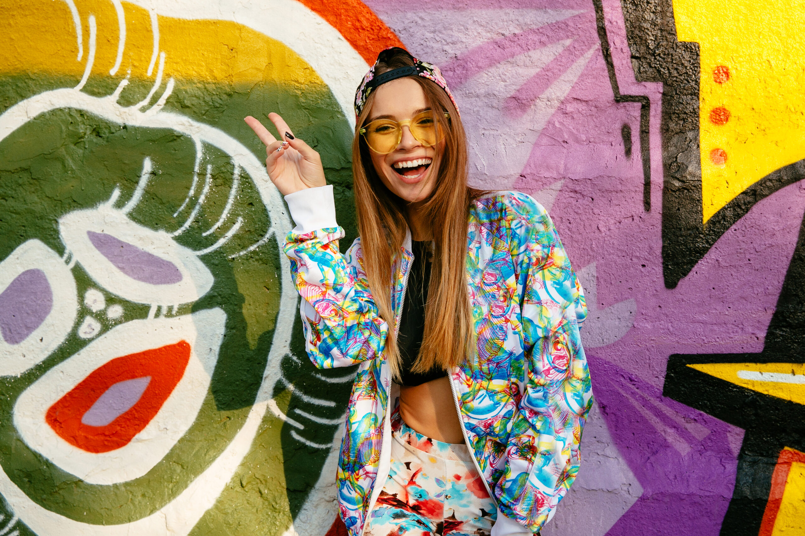 Beautiful cute girl in eyeglasses, showing peace gesture, looking at camera, widely smiling. Dressed in bright stylish clothes and cap. Posing near graffiti wall.