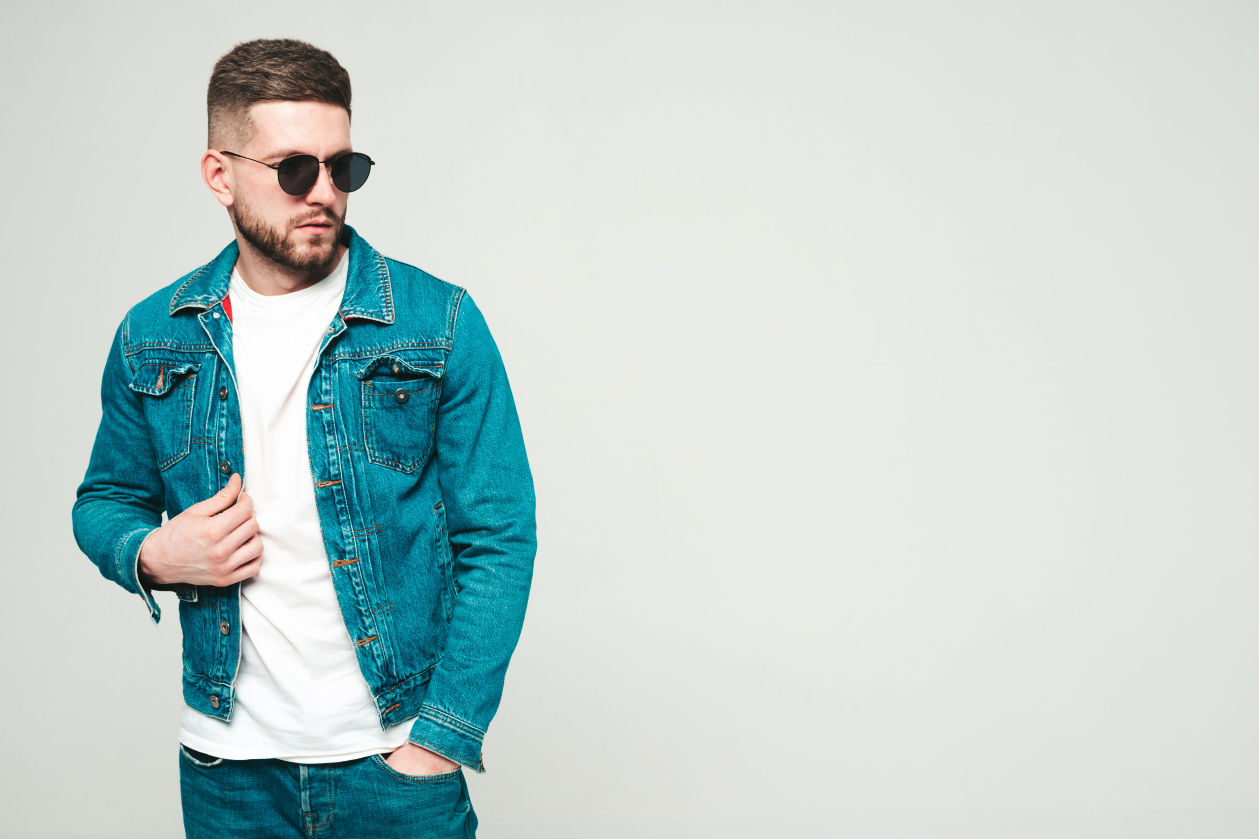 Portrait of handsome smiling stylish hipster lambersexual model.Man dressed in jacket and jeans clothes. Fashion male posing on grey background in studio in sunglasses